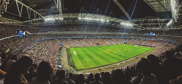 Ein Panoramablick auf ein großes, voll besetztes Fußballstadion bei Nacht, das ein laufendes Spiel auf dem hell erleuchteten Spielfeld zeigt. Die Tribünen sind mit Zuschauern gefüllt und digitale Bildschirme zeigen Spielinformationen für BelogoSports an. Das Stadiondach ist teilweise sichtbar und wölbt sich darüber.