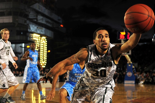 Ein Basketballspieler in weißer Camouflage-Uniform streckt mit konzentriertem Gesichtsausdruck seinen Arm aus, um einen Ball zu fangen. Hinter ihm sind weitere Spieler in blau-weißen Uniformen auf dem Platz zu sehen. Im Hintergrund sind eine Anzeigetafel und eine Industrieanlage zu sehen, die die Intensität von Basketball-Sportwetten einfangen.