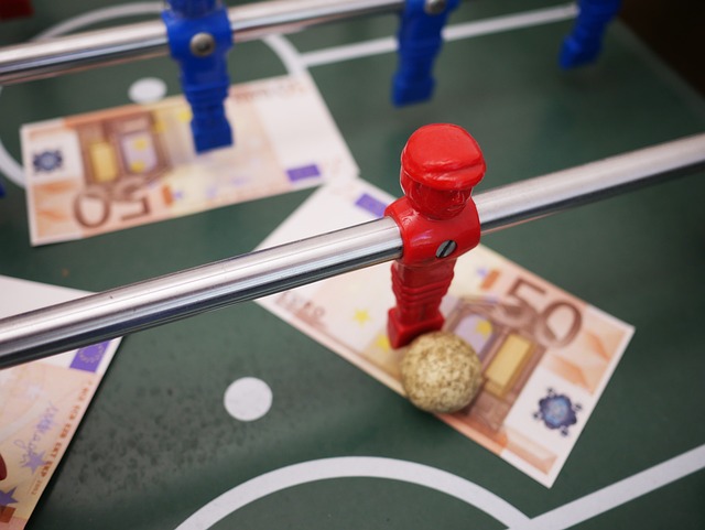 Auf einem Tisch ist ein Tischfußball-Setup mit roten und blauen Spielern zu sehen. Unter den Spielern liegen mehrere 50-Euro-Scheine, und neben einem roten Spieler steht ein Kork-Tischfußball. Die Tischoberfläche ist grün mit weißen Markierungen, was die Spannung der Bwin-Bonuscodes für neue Spieler weckt.